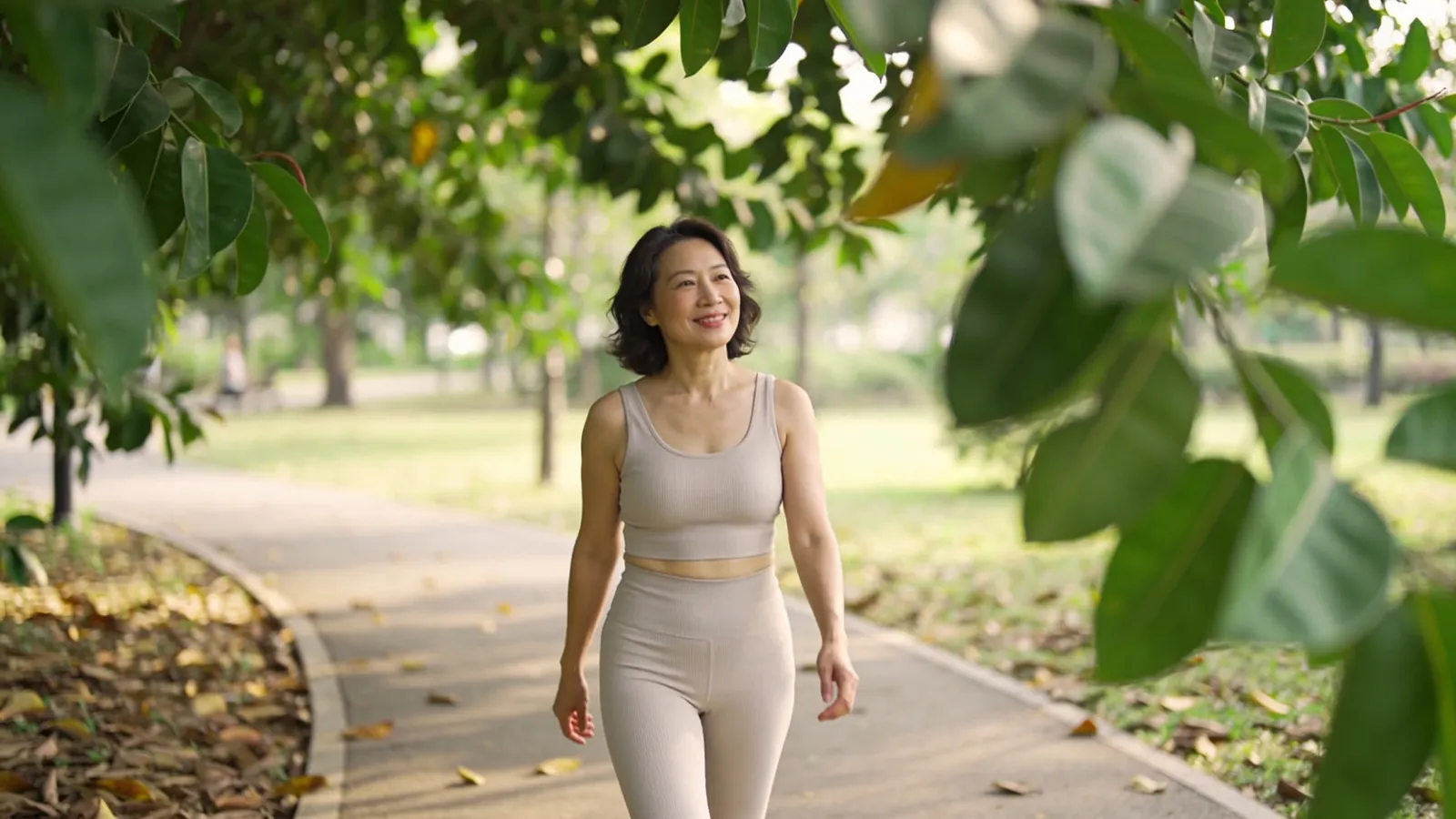East Asian woman in her forties in activewear walking along a leafy park path — illustrative image, not a patient