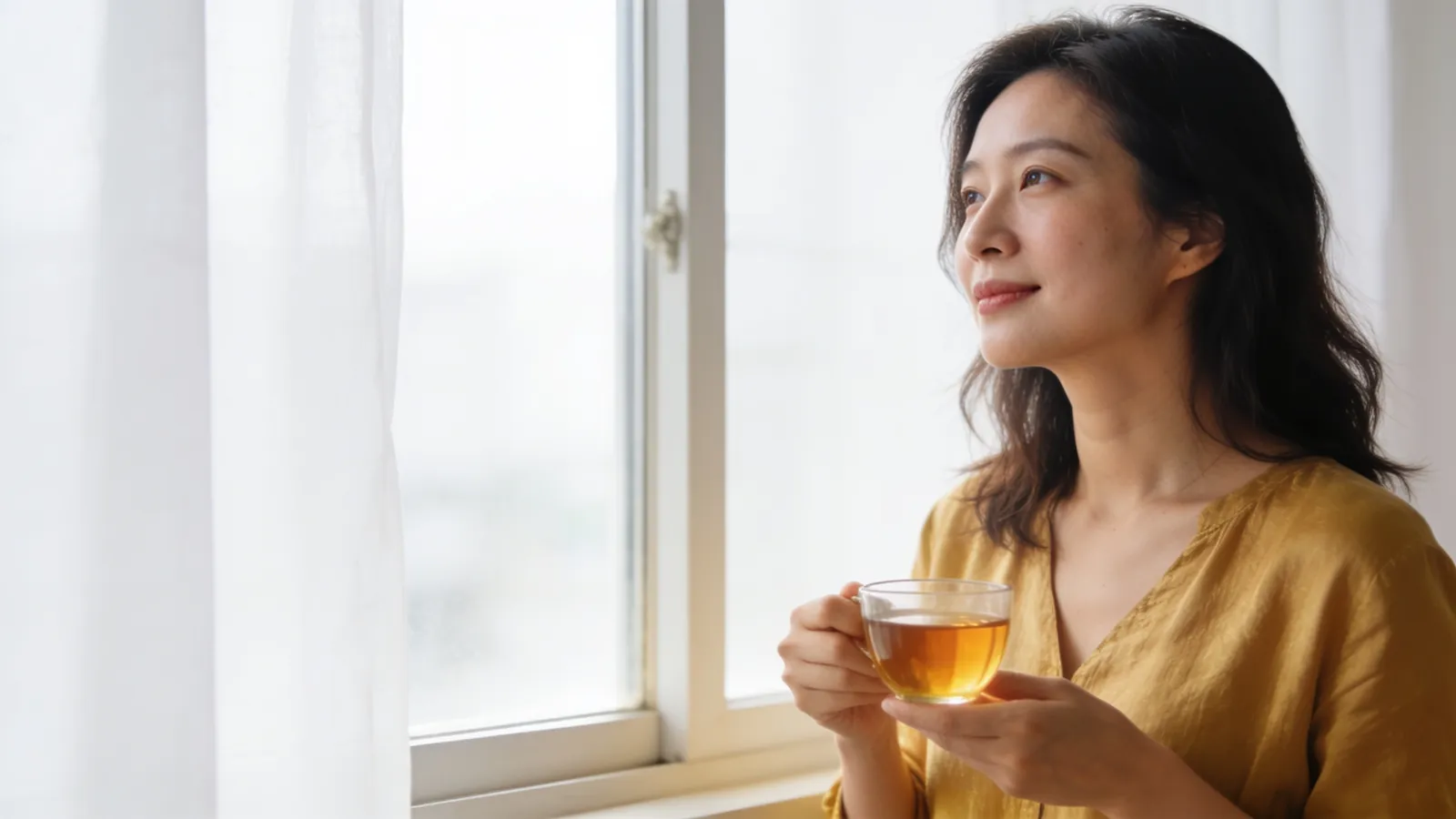 East Asian woman in her thirties holding a cup of tea and looking out of a window at home &mdash; illustrative image, not a patient